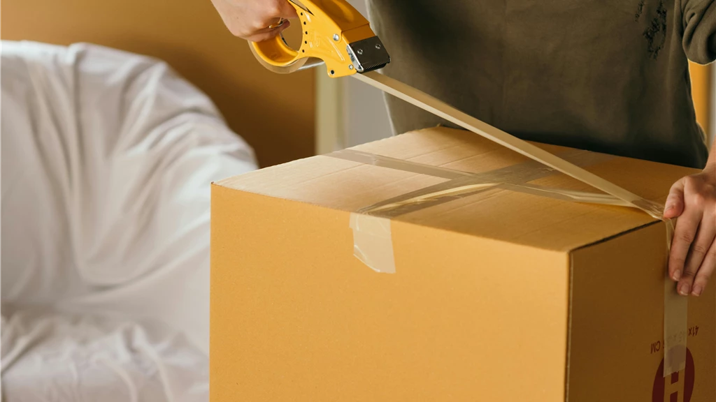 Crop female wearing casual clothes standing while packing carton boxes with scotch tape in modern apartment during preparing for relocation
