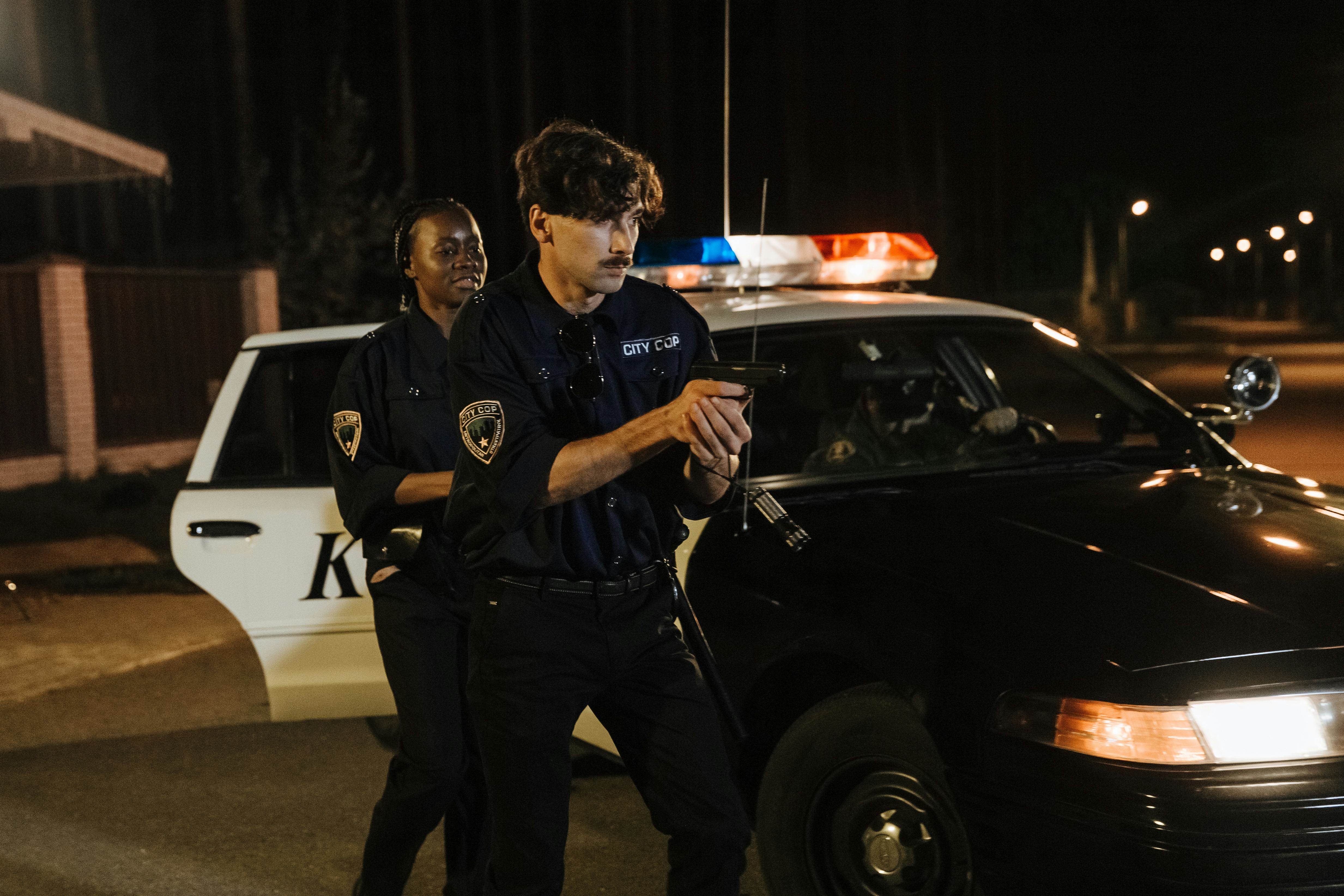 Police officers engaged in nighttime operation with a patrol car on a dimly lit street.