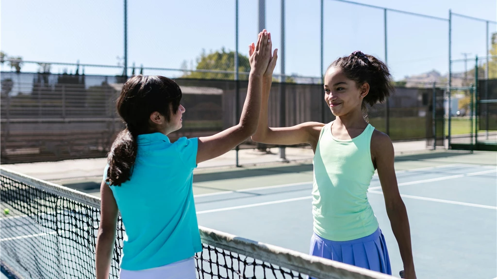 Two teen girls celebrating with a high five on a sunny tennis court.