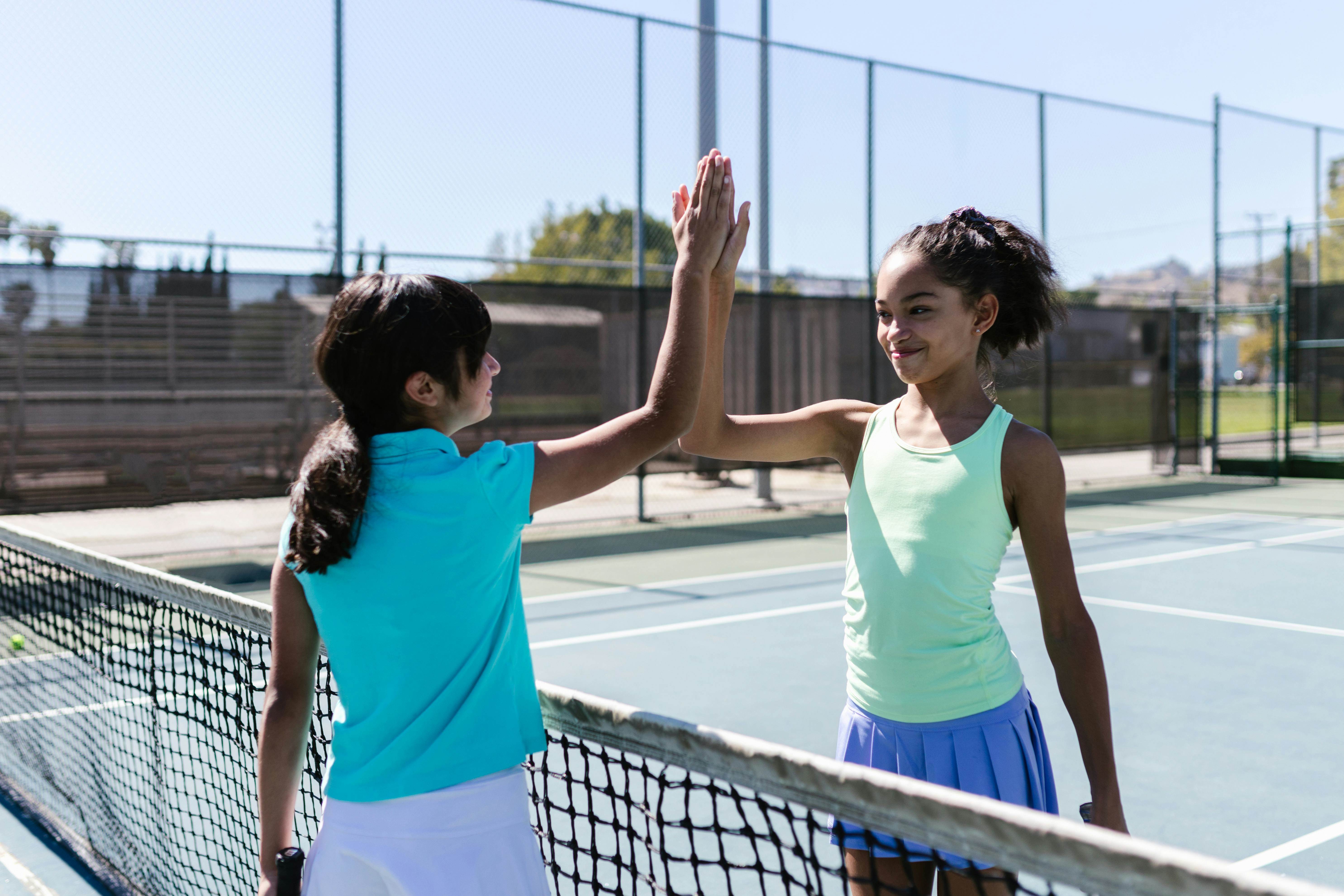 Two teen girls celebrating with a high five on a sunny tennis court.